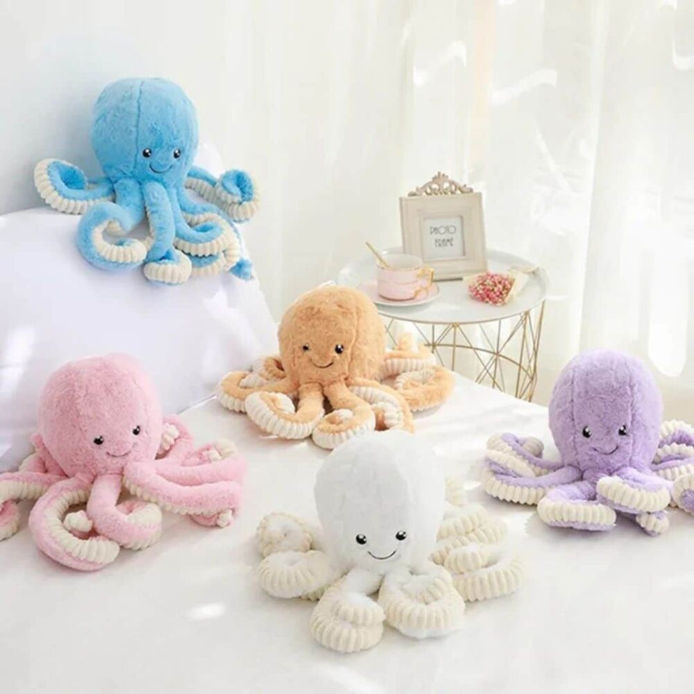 Doudou Pieuvre Mignon pour Enfant : Ensemble de cinq doudous pieuvres colorées en bleu, rose, blanc, beige et violet sur un lit.