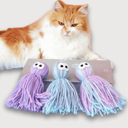 Doudou Poulpe Pour Chat 10 cm : Chat blanc et roux allongé à côté de trois doudous poulpes en laine violette et bleue.
