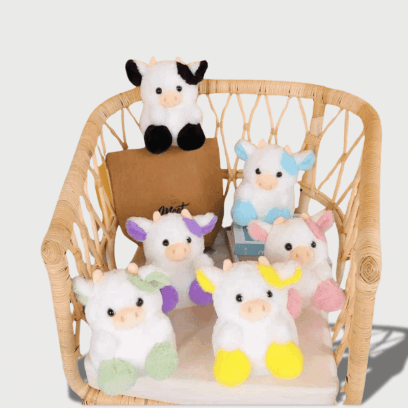 Doudou Vache Mini 20 cm : Six petits doudous vaches aux coloris variés, bleu, rose, noir, violet, jaune et gris, assis sur une chaise en osier.