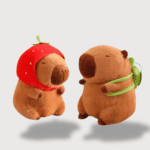 Doudou Kawaii Capybara Déguisé : Deux doudous Capybara marron, l’un avec un bonnet en forme de fraise rouge et l’autre avec un sac à dos vert.